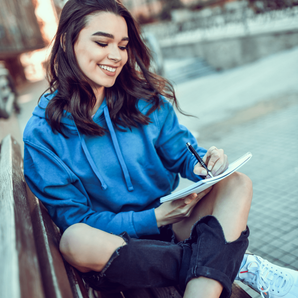 Girl sitting on a city bench writing in her notebook. She is wearing a blue sweater and jeans with the knees ripped out. This image highlights this feminist group's blog.