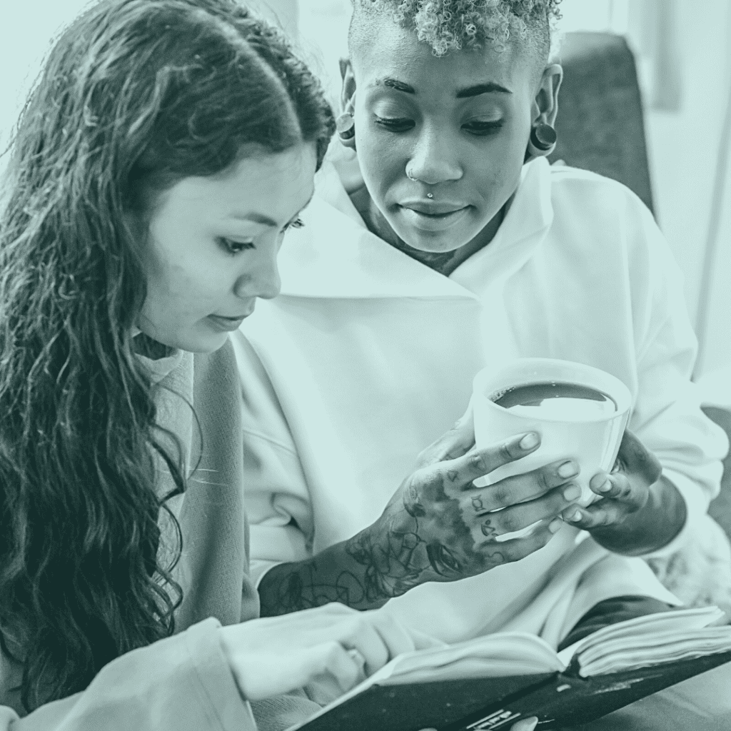 Two girls, one feminine, one looks trans look at a book/journal together while one drinks coffee.