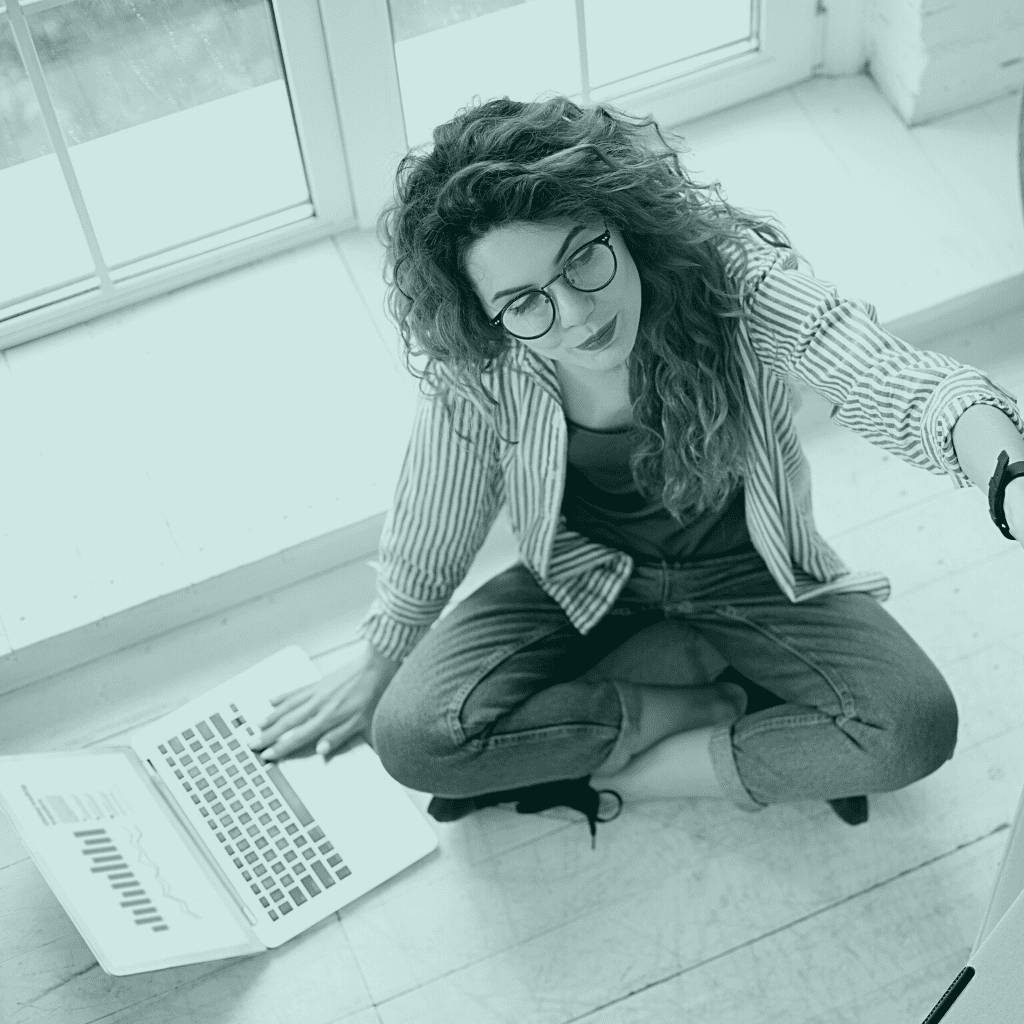 girl with curly hair sitting on the floor with a computer that has graphs on it. She is in a stripped shirt, jeans and wears glasses